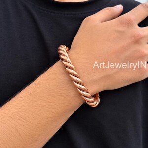 Könnte beinhalten: Ein kupferfarbenes Metallarmband mit einem gedrehten Seildesign und polierter Oberfläche. Das Armband weist eine durchgehende Spiralstruktur auf, die sich um das Handgelenk legt. Der Text ArtJewelryIN ist auf der rechten Seite zu sehen.