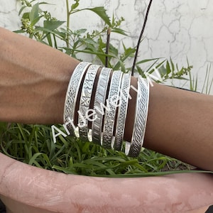 May include: A set of seven silver bangles with intricate embossed designs. The bangles are stacked on a person's wrist.