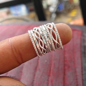 Puede incluir: Un conjunto de siete anillos de color plateado usados juntos en un dedo. La colección incluye anillos con texturas de cuerda trenzada, bandas lisas pulidas y diseños de bucle de infinito abierto, hechos de metal brillante y reflectante.