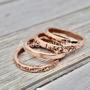 Op de afbeelding: Vier roségouden ringen met een getextureerd oppervlak zijn gestapeld op een licht houten oppervlak. De ringen hebben verschillende patronen, waaronder gehamerde en geometrische ontwerpen. De ringen hebben een warme, metallic kleur.