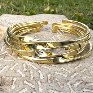 May include: A stack of gold-colored metal bracelets. Each bracelet has a twisted design and an open cuff. The bracelets are displayed on a textured, light-colored surface, with green foliage in the background.