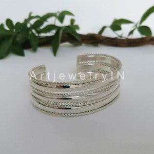 Op de afbeelding: Een zilveren manchetarmband met een gelaagd ontwerp. De armband heeft meerdere banden met een combinatie van gladde en touwachtige texturen. De tekst "ArtjewelryIN" is zichtbaar.