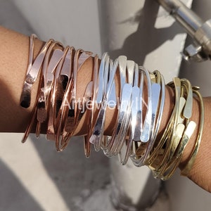 Op de afbeelding: Een verzameling gehamerde metalen armbanden in koper-, zilver- en goudtinten. De armbanden hebben een minimalistisch ontwerp met een plat, getextureerd oppervlak en een gebogen, open vorm. De armbanden zijn op een pols gestapeld.