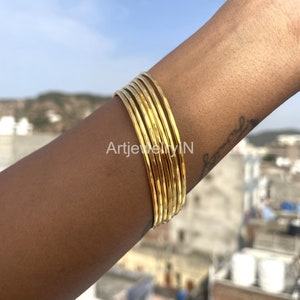 Peut inclure: Un ensemble de bracelets en métal martelé de couleur dorée. Les bracelets sont empilés sur le poignet d'une personne.