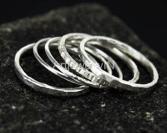 Könnte beinhalten: Eine Sammlung von fünf Silberringen mit unterschiedlichen Texturen, darunter gehämmert und geriffelt. Die Ringe sind gestapelt und zeigen ihre individuellen Muster. Die Ringe werden auf einer dunklen, strukturierten Oberfläche präsentiert.
