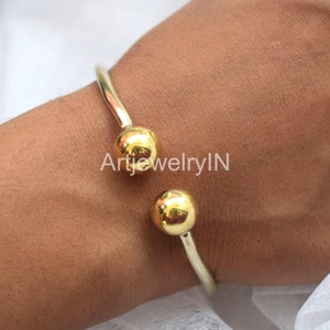 Op de afbeelding: Een gouden armband met twee ronde, gladde gouden ballen aan de uiteinden. ArtjewelryIN is op de armband gedrukt.