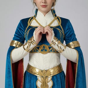 May include: A blue velvet cosplay costume with gold accents. The costume features a high collar, long sleeves, and a gold belt with a central emblem. The sleeves have a red lining, and the bodice is white with a detailed pattern.