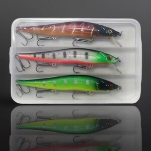 May include: A clear plastic tackle box containing three fishing lures. The lures are various colours, including brown, silver, and green, with black accents and silver hooks. Each lure has a clear plastic lip.