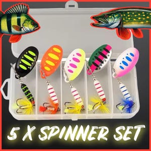 Puede incluir: Un conjunto de cinco señuelos de pesca en una caja de plástico transparente. Los señuelos son de diferentes colores y tienen una variedad de diseños. La caja tiene una etiqueta que dice "5 x Spinner Set".