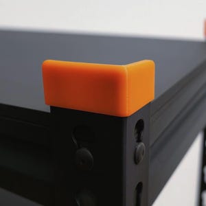 Könnte beinhalten: Ein schwarzes Metallregal mit leuchtend orangefarbenen L-förmigen Eckschonern, die an den oberen Kanten der vertikalen Stützpfosten angebracht sind. Das Regal hat eine mattschwarze Oberfläche mit sichtbaren vertikalen Schlitzen am Rahmen.