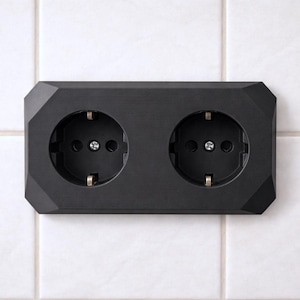 Könnte beinhalten: Eine schwarze Doppelsteckdose mit geometrischem Design. Die Steckdose hat zwei runde Buchsen und ist an einer weiß gefliesten Wand montiert. Die Buchsen haben Metallkontakte und sind im schwarzen Rahmen zentriert.