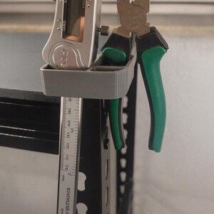Könnte beinhalten: Ein grauer, 3D-gedruckter Werkzeughalter an einem Metallregal. Er hält einen digitalen Messschieber und eine Zange mit grünen Griffen. Ein Edelstahl-Lineal mit Zentimetermessung ist ebenfalls sichtbar.