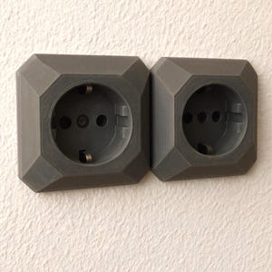 Puede incluir: Dos enchufes eléctricos de color gris oscuro con un diseño octogonal están montados en una pared blanca texturizada. Cada enchufe tiene tres agujeros circulares y dos clavijas metálicas. Los enchufes están uno al lado del otro.
