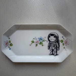 Può includere: Vassoio rettangolare bianco in ceramica con un'illustrazione di un astronauta rana. Il vassoio presenta accenti floreali rosa e blu. La rana astronauta indossa una tuta spaziale. Il vassoio ha una forma ottagonale.