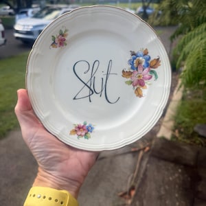 Può includere: Piatto in ceramica bianca con bordo dorato e decorazioni floreali. La parola "Shit" è scritta in corsivo nero al centro del piatto. Il piatto ha un bordo smerlato.