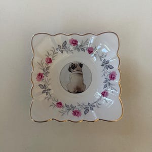 May include: A white, square decorative dish with a scalloped edge and gold trim. The dish features a central image of a pug dog and is surrounded by a floral design with pink roses and grey leaves. A charming accent piece.