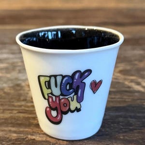 May include: A white ceramic espresso cup with a black interior. The exterior features the text "FUCK YOU" in a colourful, bubble-style font with a small red heart graphic positioned to the right of the lettering, creating a bold visual contrast.