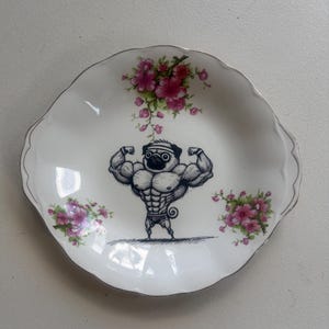 Può includere: Piatto decorativo bianco con bordo smerlato e bordo argentato. Il piatto presenta un'illustrazione di un carlino muscoloso che flette i suoi bicipiti. Accenti floreali rosa sono posti sul bordo del piatto.