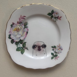 Può includere: Piatto in ceramica bianca con bordo dorato, con un'illustrazione di carlino disegnata a mano e dettagli floreali di rose rosa, bianche e verdi. Il piatto ha un bordo smerlato.