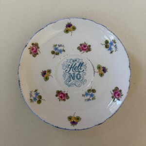 May include: A white ceramic saucer with a blue rim, decorated with a floral pattern. The centre of the saucer features the phrase "OH Hell NO" in blue script, surrounded by small pink, purple, and blue flowers. The edge has a distressed look.