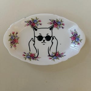 May include: White oval ceramic dish featuring a black cat wearing sunglasses and giving the middle finger. The dish is decorated with floral arrangements in pink, purple, and blue.