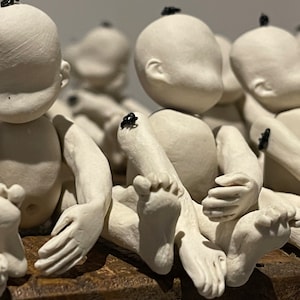 May include: A group of white ceramic baby dolls with black dots on their heads, arranged in a row. The dolls are sitting with their legs crossed and their hands clasped together.