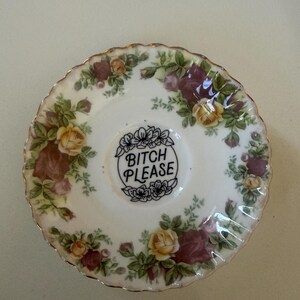 May include: A white ceramic saucer with a floral design in yellow, burgundy, and green. The centre of the saucer features the phrase "BITCH PLEASE" in black script, surrounded by a floral wreath. The rim has a gold trim.