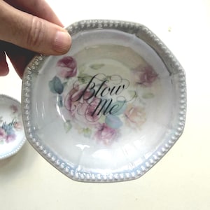 Può includere: Piccolo piatto ottagonale iridescente con bordo perlato. Il piatto presenta un motivo floreale rosa, viola e giallo, con le parole "Blow Me" in corsivo. Un secondo piatto è parzialmente visibile sullo sfondo.