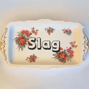 Può includere: Vassoio rettangolare color crema con un motivo floreale a rose e la parola "Slag" in grassetto nero. Il vassoio ha manici e finiture dorate.