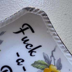 Può includere: Vassoio in ceramica bianca con un motivo floreale e le parole "Fuck -e-" in corsivo nero. Il vassoio ha un bordo dorato e un bordo decorativo. Sono visibili fiori gialli e viola.