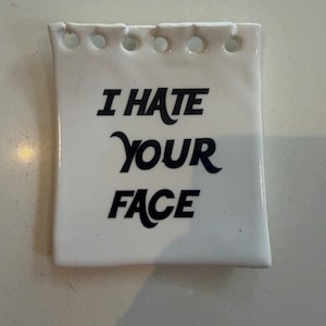 May include: A white ceramic square with rounded corners and a row of holes along the top edge. The text "I HATE YOUR FACE" is printed in black, stylized font. The item appears to be a decorative piece.
