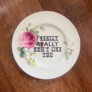 Può includere: Un piatto decorativo bianco con una rosa rosa e foglie verdi. Il piatto ha un bordo dorato e il testo "I REALLY REALLY DON'T LIKE YOU" in nero. Posizionato su una superficie di legno.