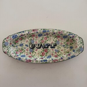 Può includere: Vassoio ovale in ceramica con un motivo floreale rosa, blu e verde. La parola "CUNT" è stampata in lettere nere e blu al centro del vassoio. Il vassoio ha un bordo smerlato.
