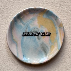 May include: A small, round, ceramic dish with a marbled design in shades of pink, blue, and yellow. The dish has the word "MINGE" written in black letters.