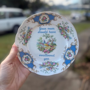 Può includere: Un piatto di porcellana bianca con un motivo floreale blu e oro. Il piatto presenta il testo "Your mum should have swallowed you" in blu. Una scena centrale raffigura uccelli e fiori.