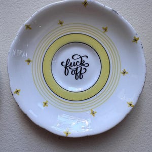 Può includere: Piattino in ceramica bianca con centro giallo e cerchi concentrici. Il centro presenta la scritta "fuck off" in corsivo nero. Piccoli accenti floreali gialli sul bordo.