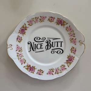 Può includere: Piatto in ceramica bianca con bordo dorato e motivo floreale. Il piatto presenta la scritta "NICE BUTT" in nero. Rose rosa e foglie verdi decorano il bordo del piatto.