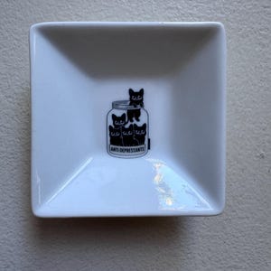 May include: A white, square ceramic dish featuring a black graphic of black cats in a jar. The jar has the words "ANTI DEPRESSANTS" printed on it. The dish has a slightly angled design.