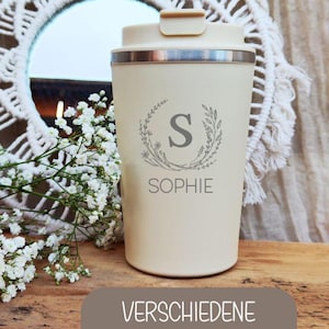 Könnte beinhalten: Ein cremefarbener Reisebecher mit silbernem Rand und Deckel. Der Becher ist mit dem Namen "SOPHIE" und einem floralen Kranzdesign um den Buchstaben "S" personalisiert. Der Text "VERSCHIEDENE MOTIVE" wird auf braunem Hintergrund angezeigt.