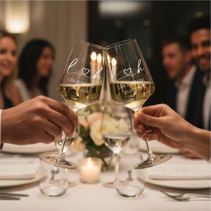 May include: Two clear glass wine glasses held in a toast, each featuring white printed text that reads L & H with a heart symbol, followed by LISA & HENRIK and the date 25.05.2026. The glasses are filled with a pale yellow liquid.