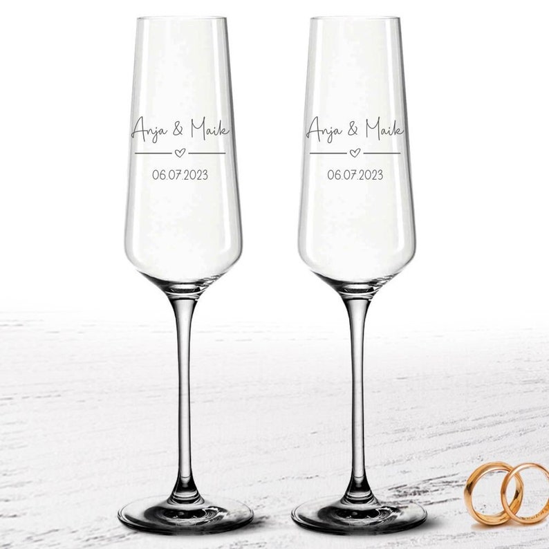 Puede incluir: Dos copas de champ&aacute;n transparentes con los nombres "Anja & Maike" y la fecha "06.07.2023", separados por un coraz&oacute;n. Dos anillos de boda dorados se ven en la esquina inferior derecha.