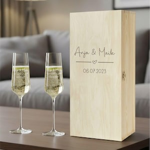 Puede incluir: Dos copas de champ&aacute;n llenas de l&iacute;quido burbujeante junto a una caja de madera. La caja est&aacute; grabada con los nombres "Anja & Maik", un coraz&oacute;n y la fecha "06.07.2023". Las copas y la caja est&aacute;n sobre una superficie marr&oacute;n.