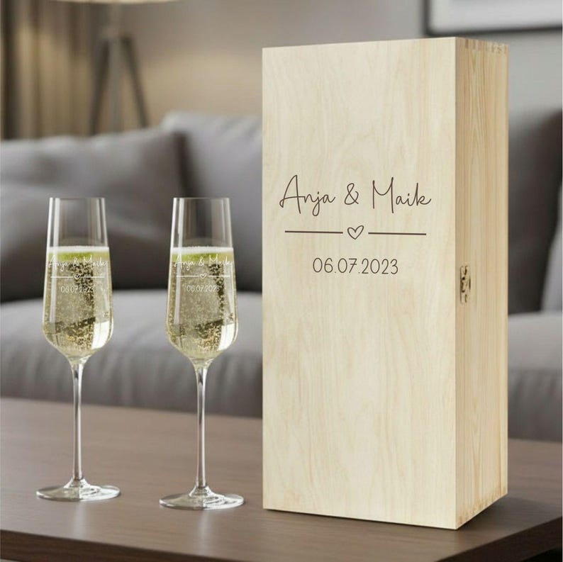 Puede incluir: Dos copas de champ&aacute;n llenas de l&iacute;quido burbujeante junto a una caja de madera. La caja est&aacute; grabada con los nombres "Anja & Maik", un coraz&oacute;n y la fecha "06.07.2023". Las copas y la caja est&aacute;n sobre una superficie marr&oacute;n.
