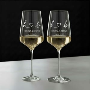 May include: Two clear glass wine glasses featuring white etched text that reads h heart b, HELENA & BERND, and 14.02.2025. The glasses have long, slender stems and are filled with a pale yellow liquid, set against a dark and light background.