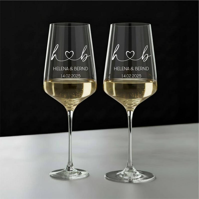 May include: Two clear glass wine glasses featuring white etched text that reads h heart b, HELENA & BERND, and 14.02.2025. The glasses have long, slender stems and are filled with a pale yellow liquid, set against a dark and light background.