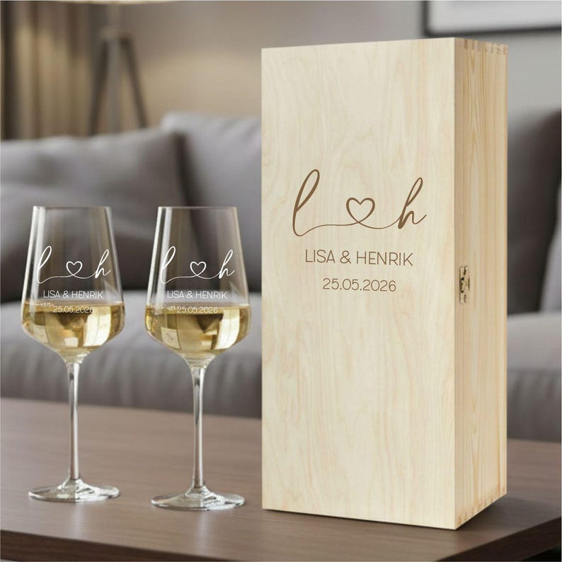 May include: Two clear glass wine glasses and a tall rectangular light-colored wooden box. Each glass and the box feature the engraved text L, a heart symbol, h, the names LISA & HENRIK, and the date 25.05.2026 in a matching script font.