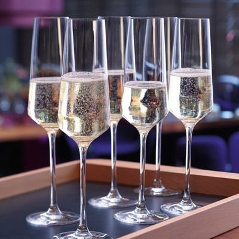 Puede incluir: Seis copas de champ&aacute;n transparentes llenas de vino espumoso, dispuestas sobre una bandeja de madera. Las copas tienen tallos largos y delgados y un dise&ntilde;o de cuenco angular. El vino es de color amarillo p&aacute;lido, con burbujas visibles.