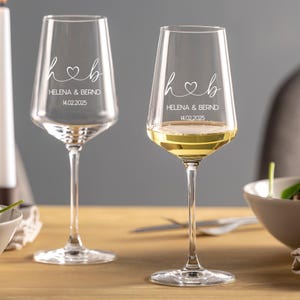 May include: Two clear glass wine glasses with long stems. Each glass features white etched text reading h heart b, HELENA & BERND, and 14.02.2025. One glass is empty, while the other contains a pale yellow liquid, showcasing the custom engraving.