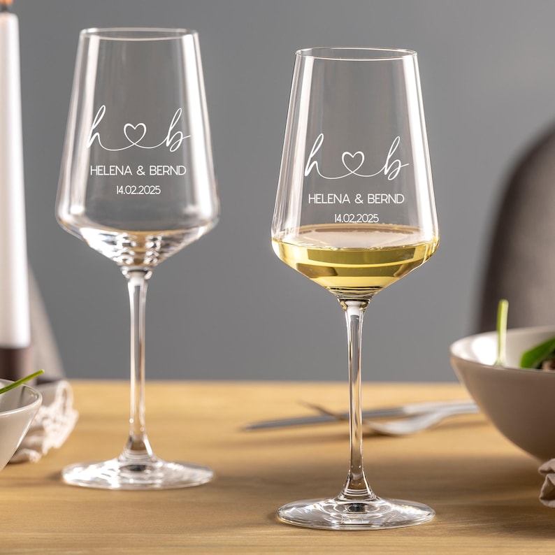 May include: Two clear glass wine glasses with long stems. Each glass features white etched text reading h heart b, HELENA & BERND, and 14.02.2025. One glass is empty, while the other contains a pale yellow liquid, showcasing the custom engraving.