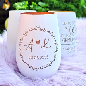 Könnte beinhalten: Zwei weiße Glas-Kerzenhalter mit goldfarbenem Rand. Der Kerzenhalter links hat ein florales Kranz-Design mit den Initialen "A & K" und dem Datum "20.05.2023". Der Kerzenhalter rechts hat den Text "Der schönste Weg ist der gemeinsame" und "Johanna & S 15.07.2023".